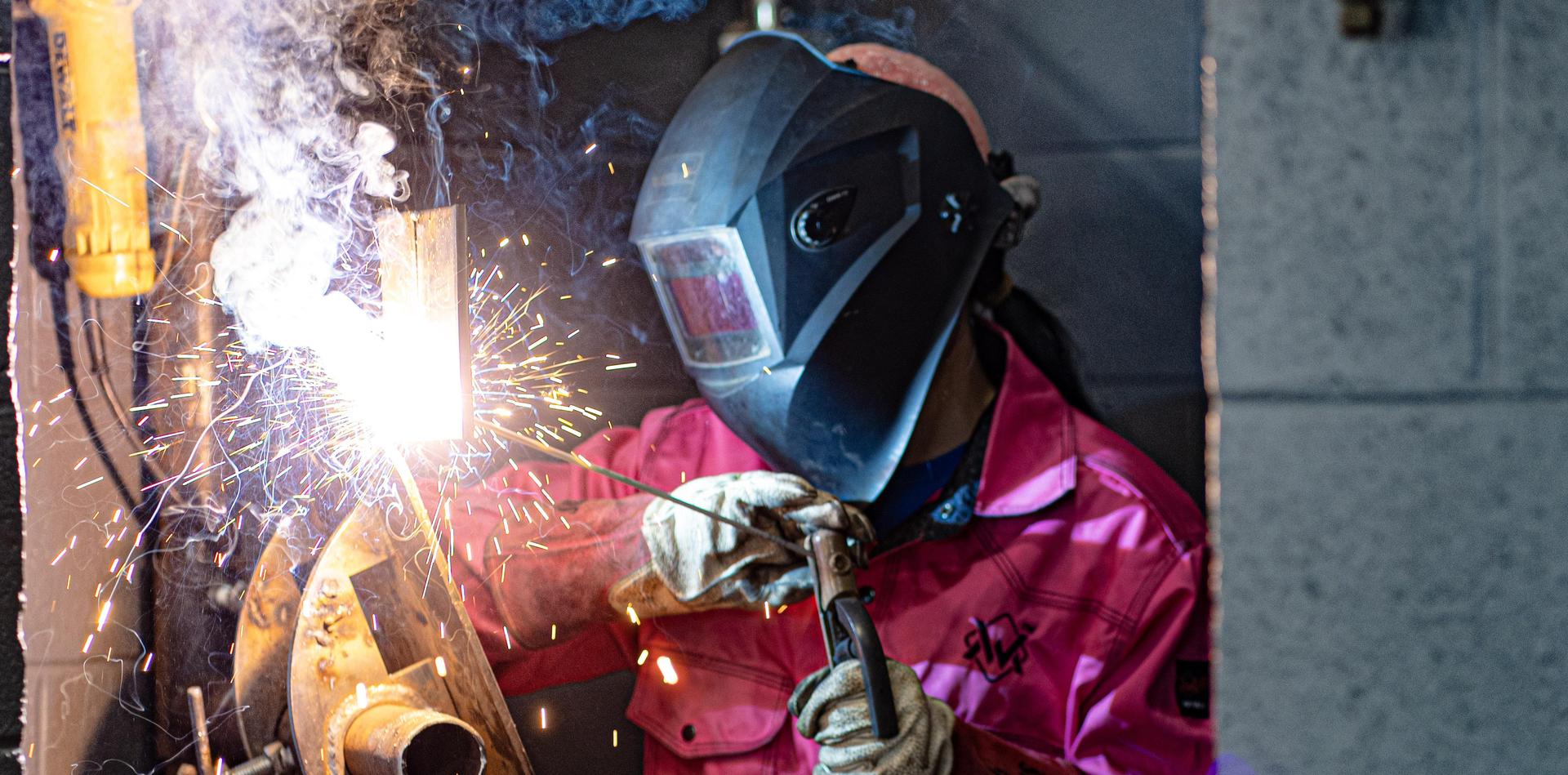 Welder in a pink shirt welding metal, sparks flying in a workshop setting.