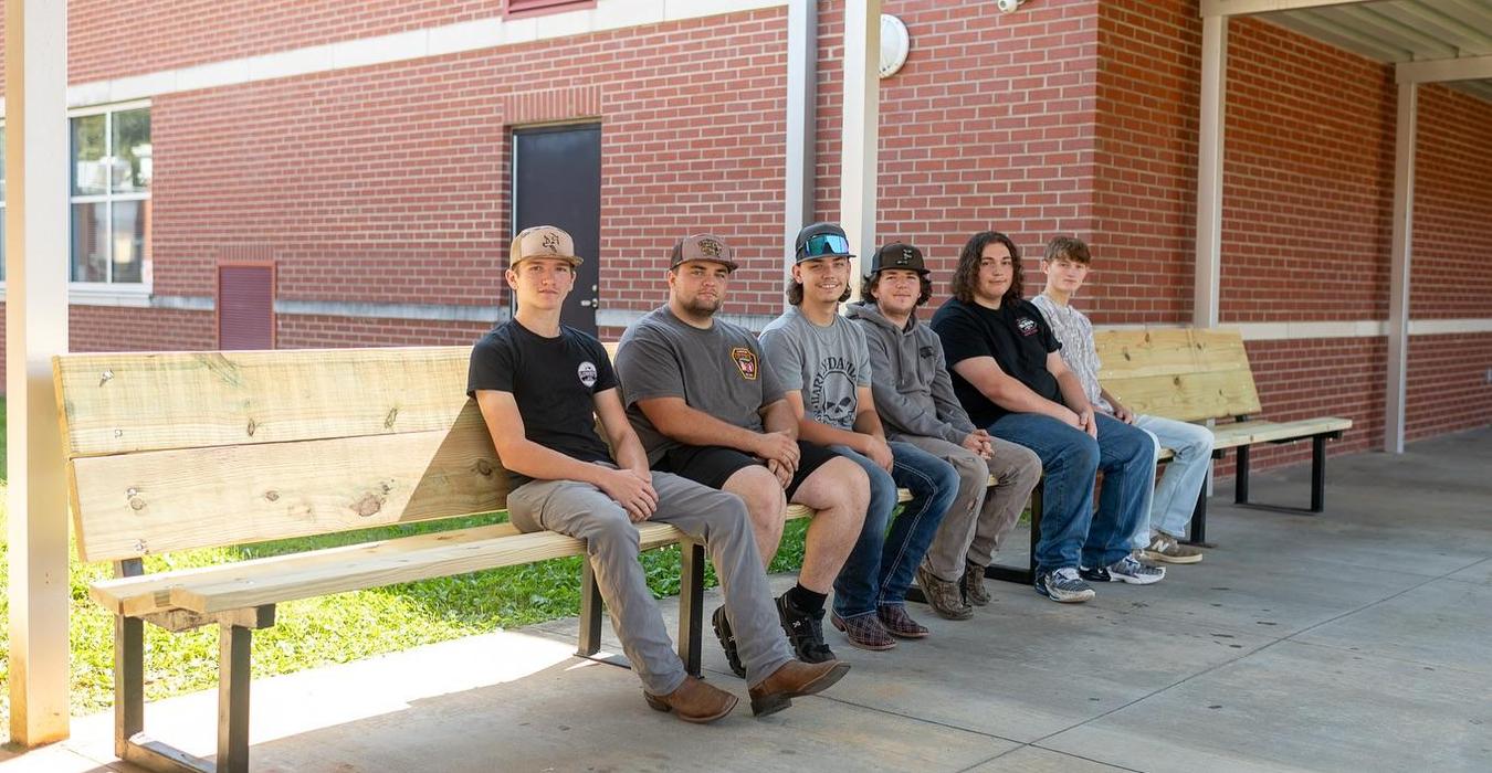 Six male students sitting on benches outside a school building.