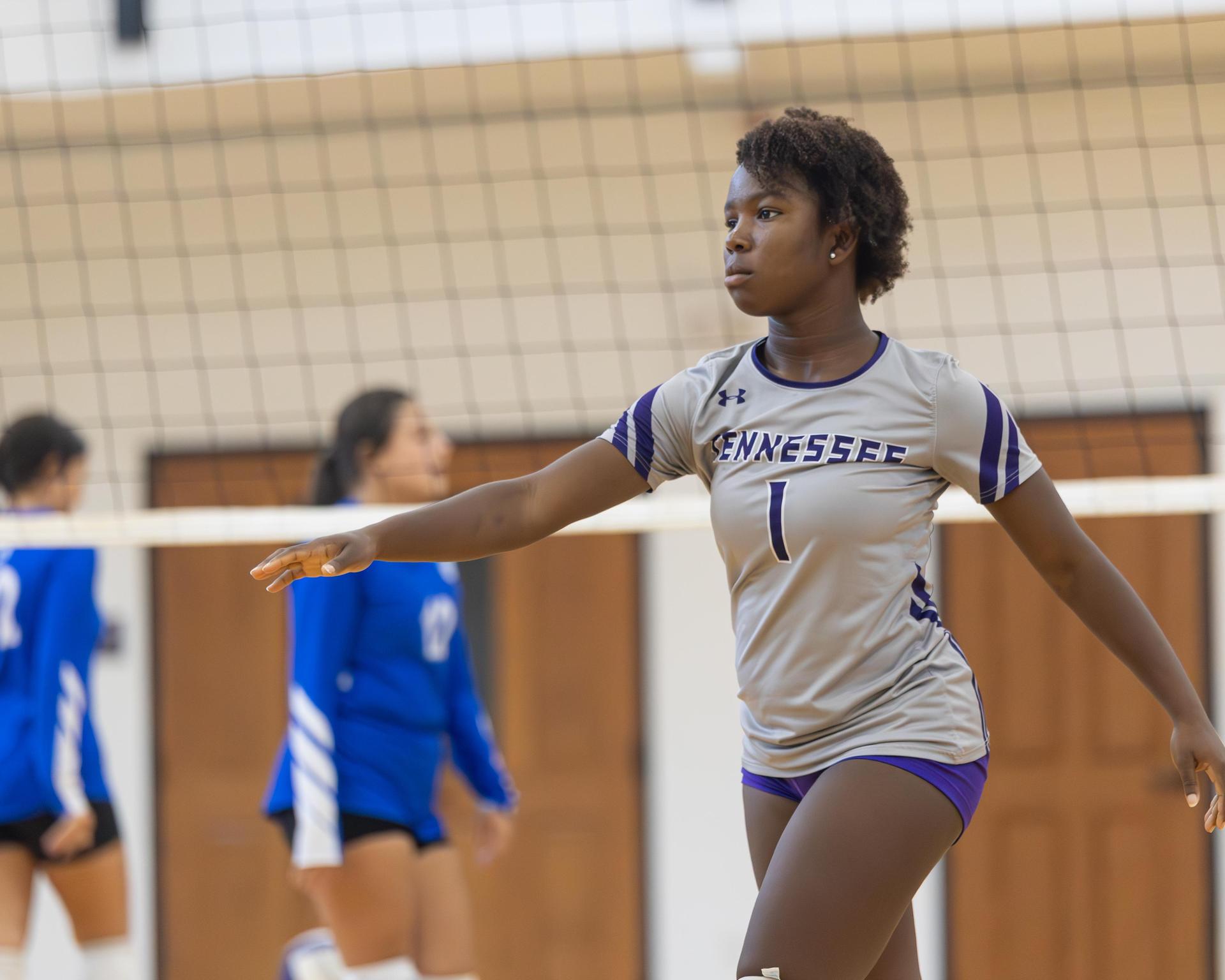 A volleyball player in gray stands in a gym, signaling during a game.