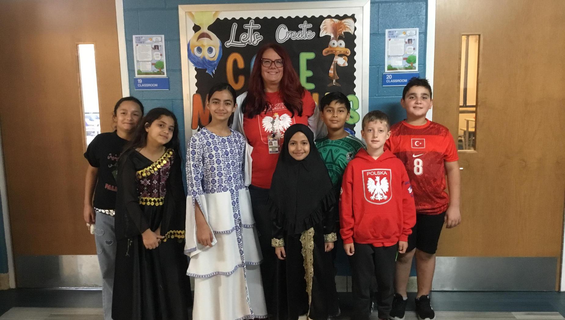 Group of kids with a teacher, dressed in various cultural outfits, in a school hallway.