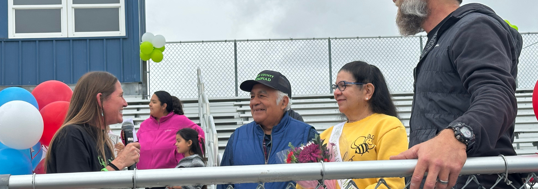 A man and woman smile while receiving flowers during a community event with balloons.