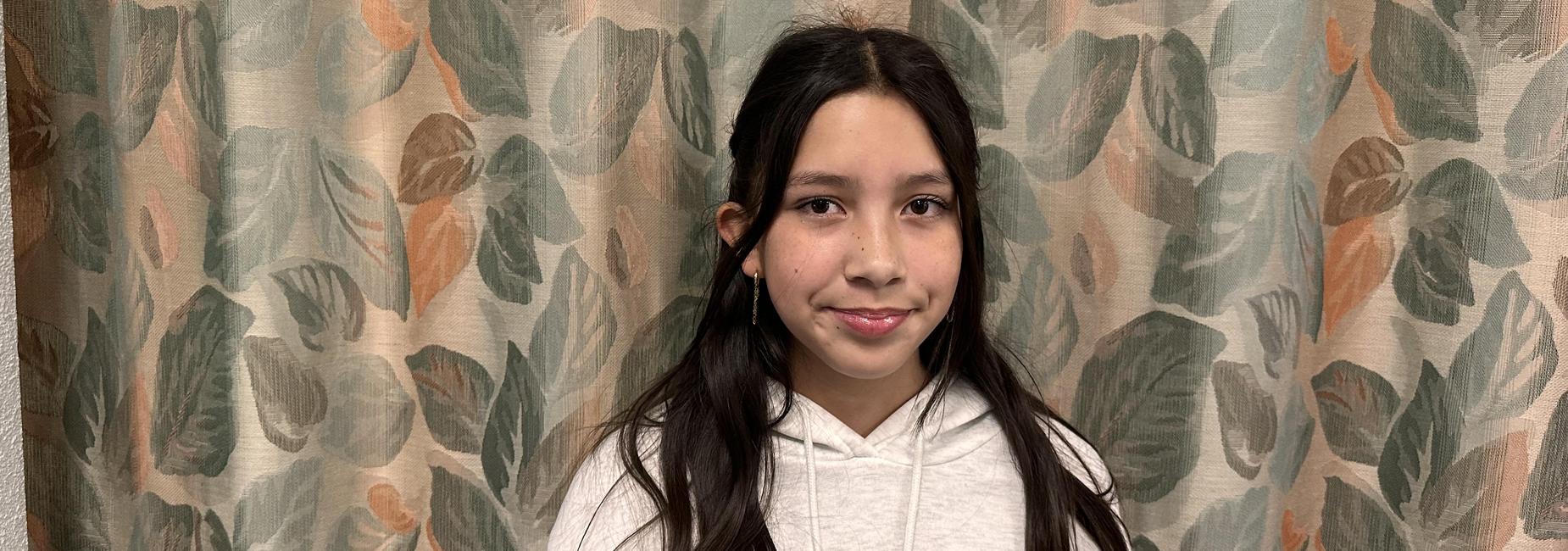 Young girl in a white hoodie standing in front of a leafy patterned curtain.