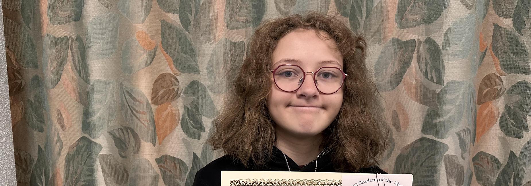 Girl holding a certificate for Student of the Month, smiling in front of patterned curtains.
