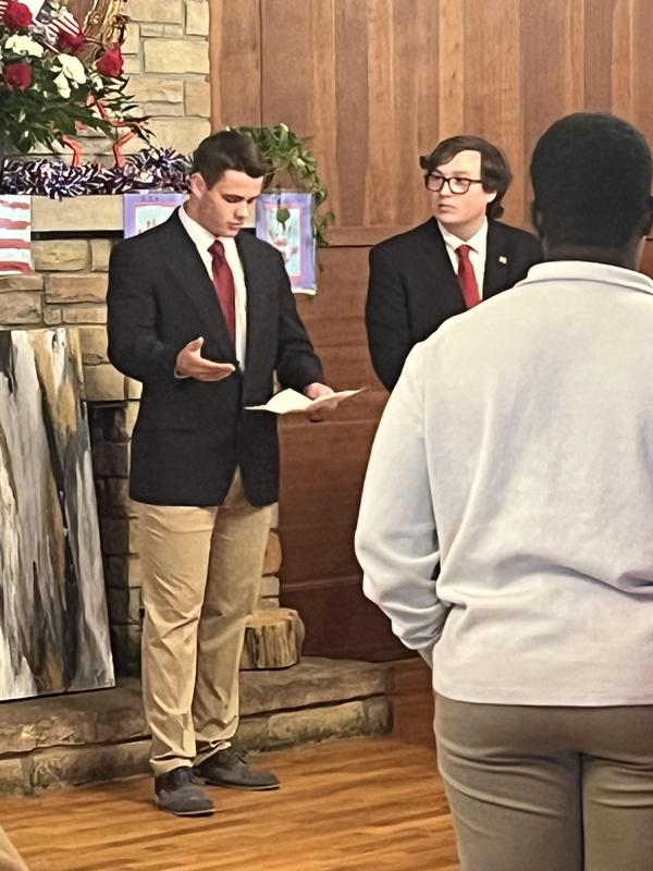 Rylen speaks before the group assembled in the Community House with Zach looking on.
