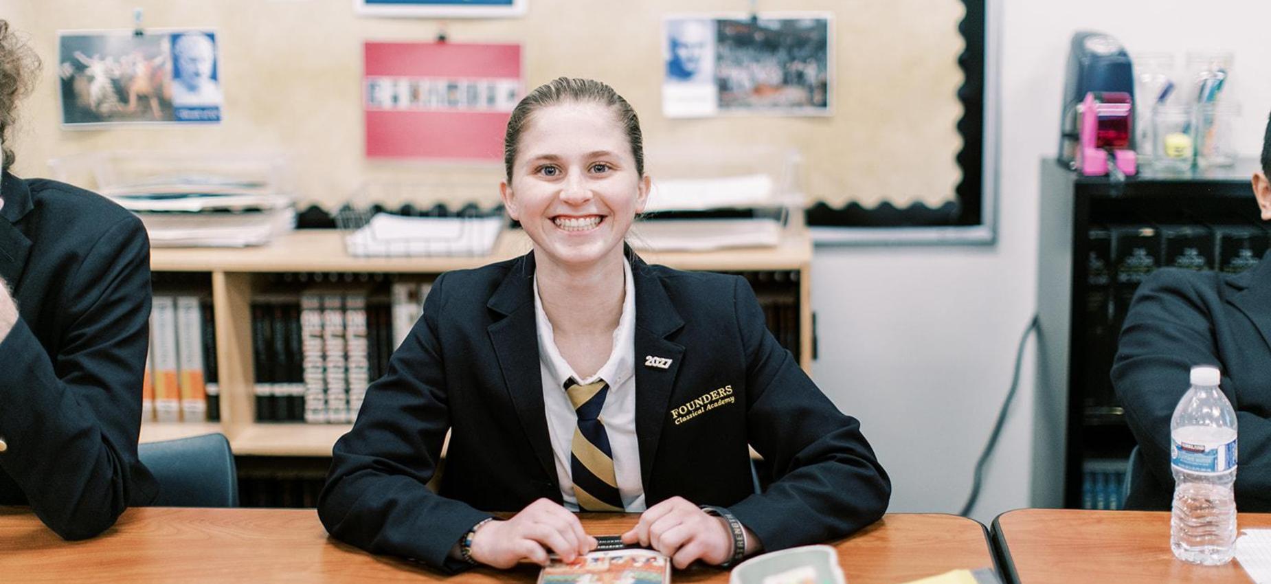 Smiling high school student in a navy blazer sits at her desk