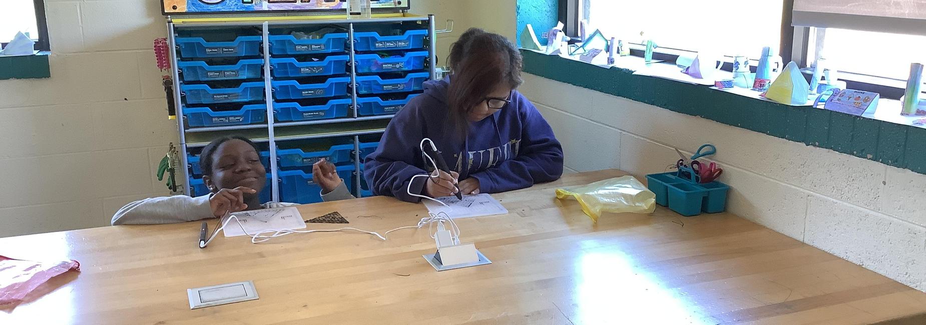 Two children working at a table, one drawing while the other smiles.