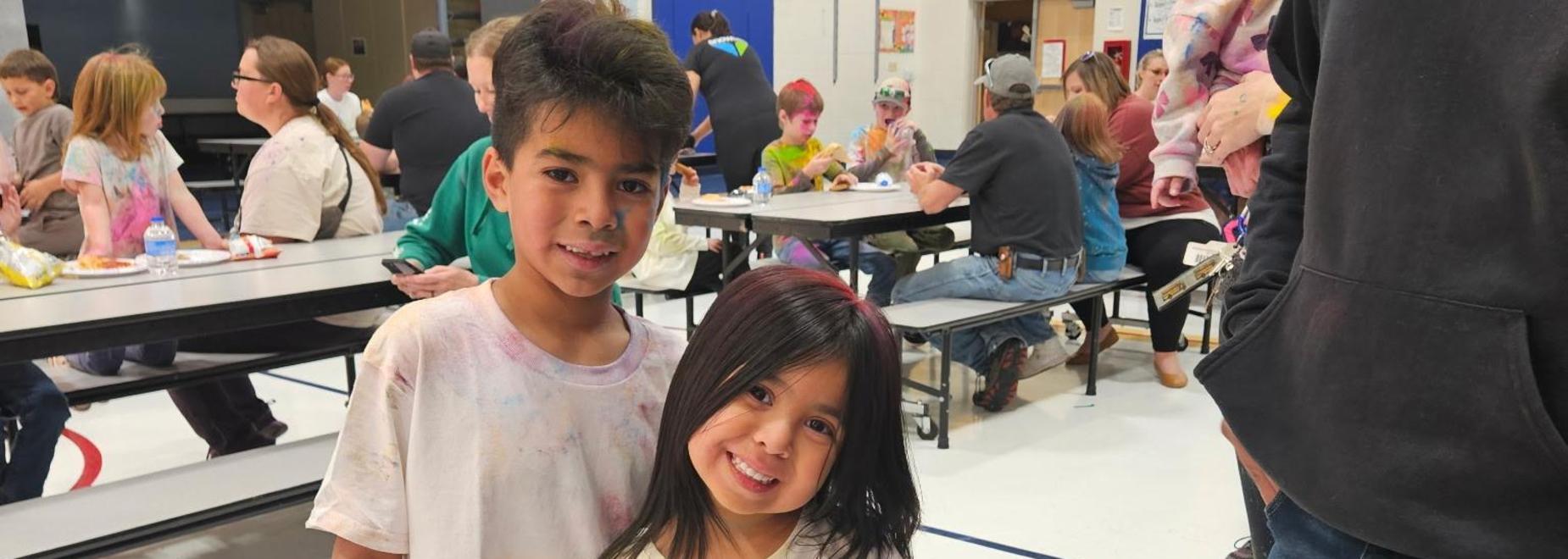 Two children smiling in a school cafeteria filled with people.