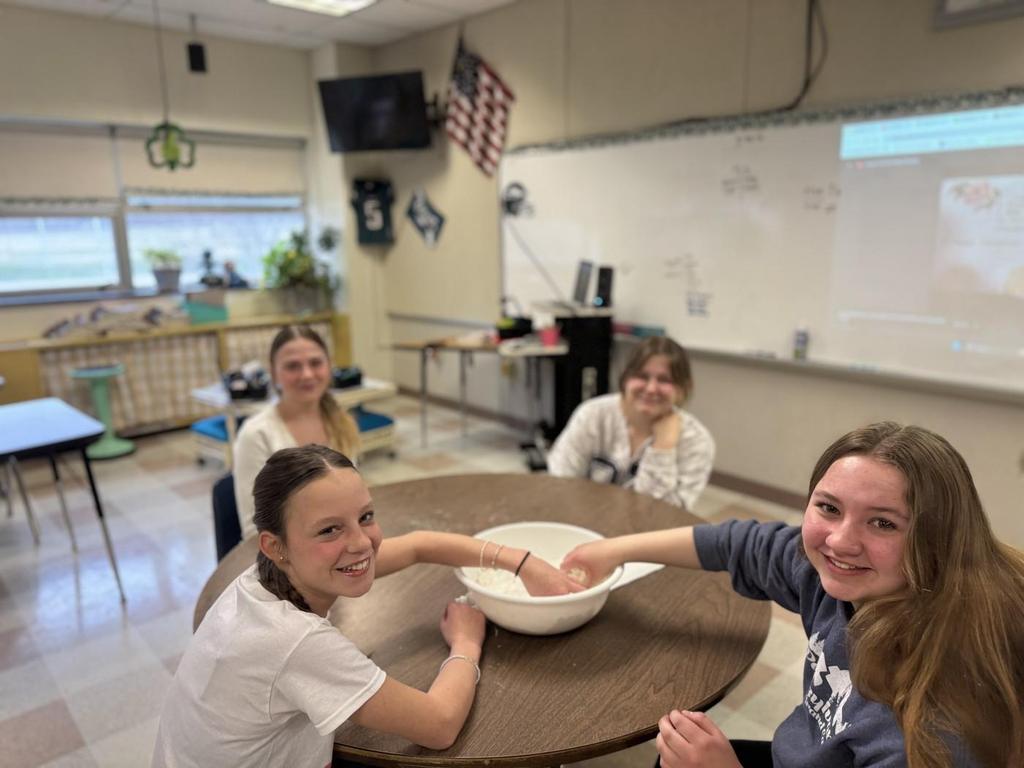 Four students sitting at a table in a classroom -- two students have their hands in a bowl that is on the table.
