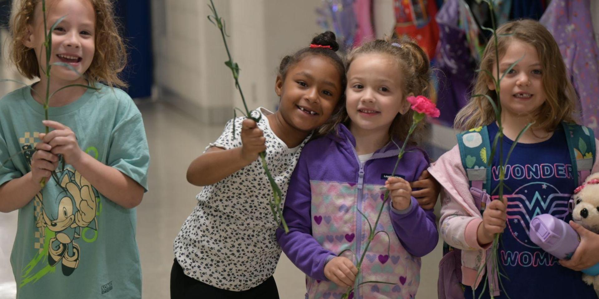 Four children holding flowers in a hallway, smiling at the camera.