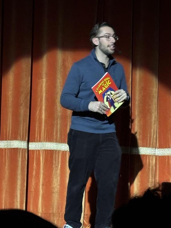 Male performer in a blue sweater holding a magic book on stage.