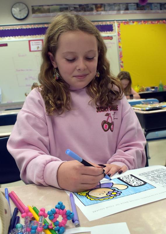 Girl coloring at a desk with other students in the background.