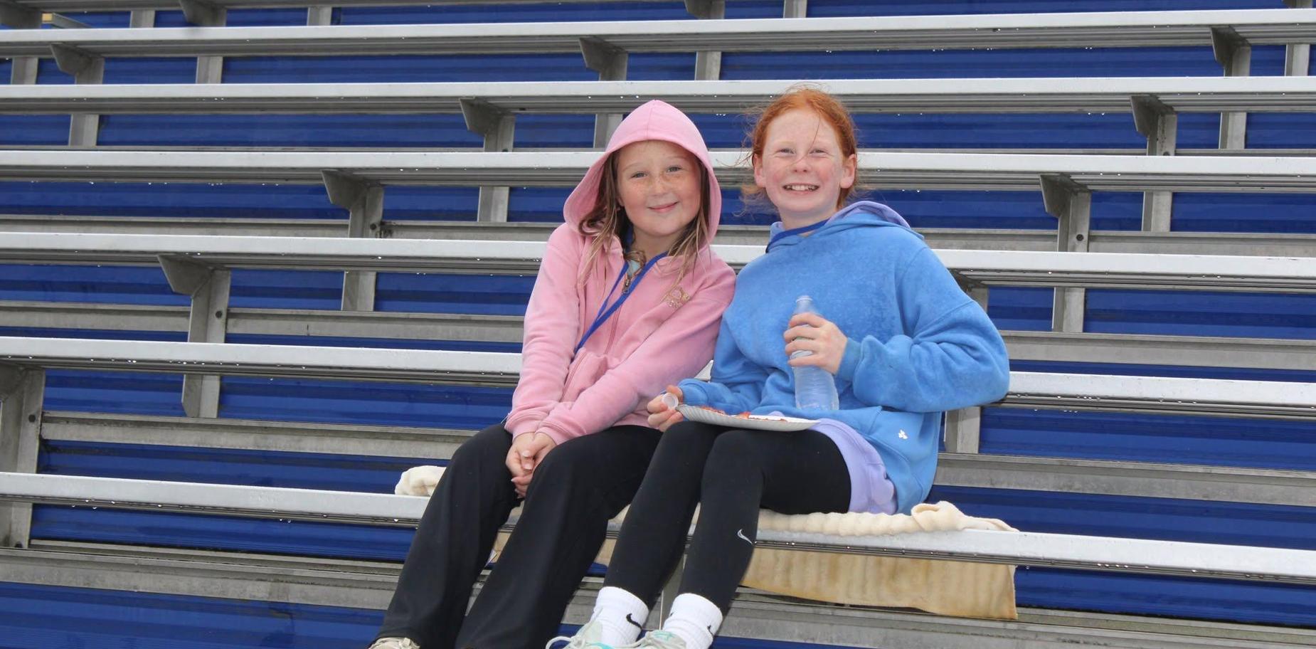Two girls sit on bleachers, smiling while enjoying snacks and drinks.