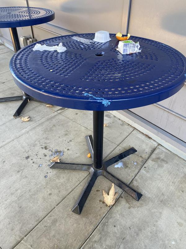 Round blue metal outdoor table with perforated surface, littered with empty food containers, napkins, and an orange peel. A