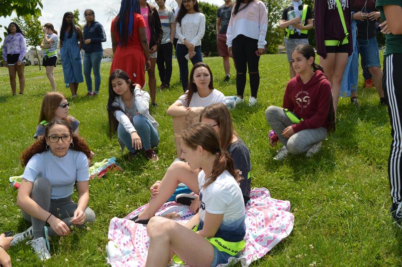 7 girls (three 6th graders and three 0th graders) are sitting on the grass. A group are standing behind waiting to meet their buddies