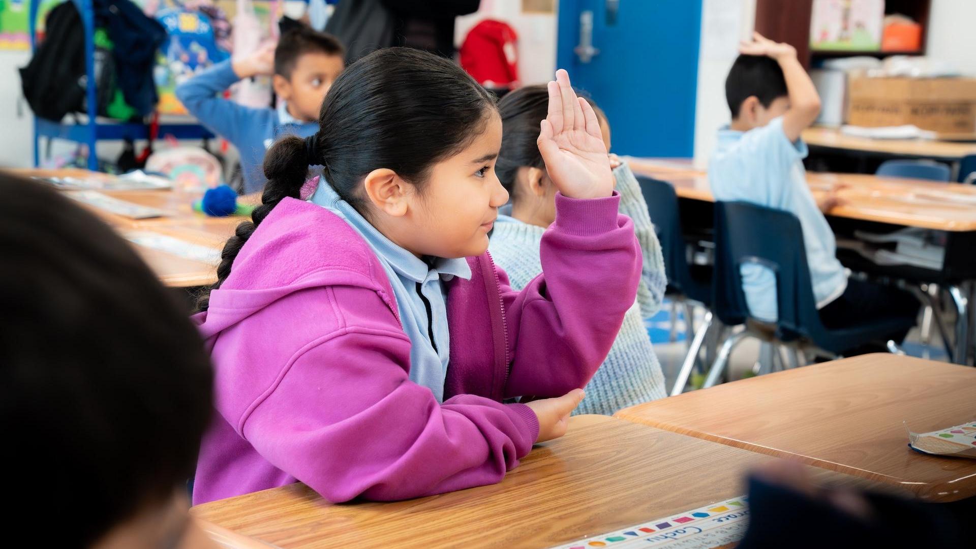 A girl in a purple jacket raises her hand in a classroom filled with students.