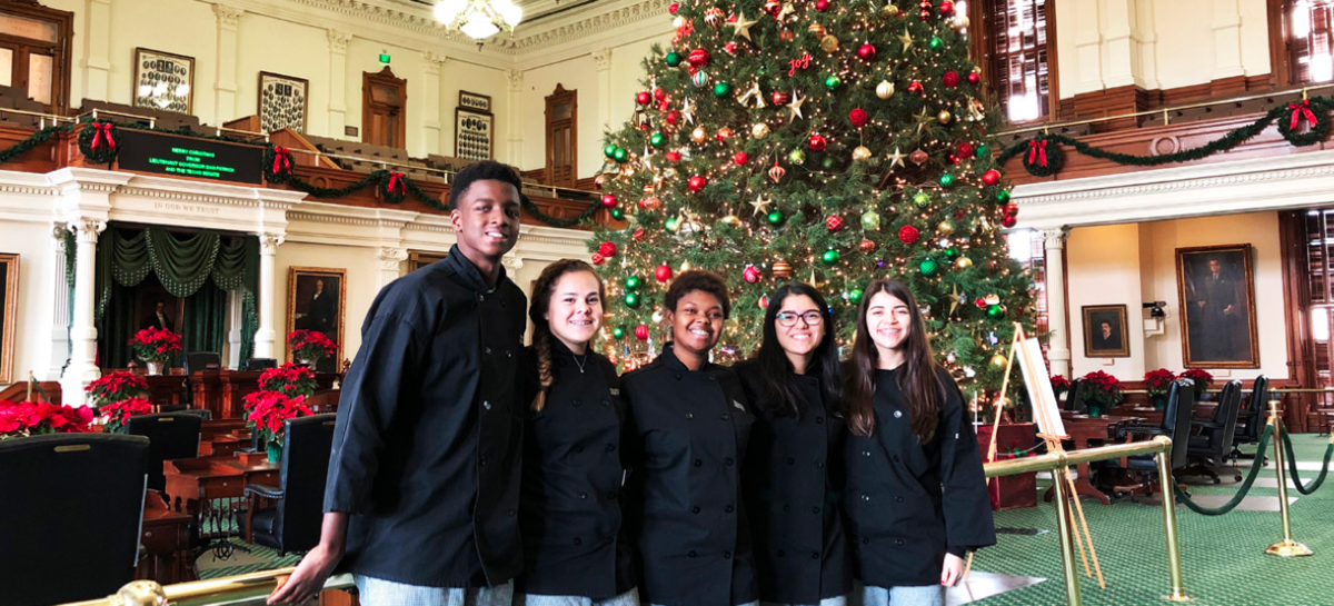 Texas School for the Deaf (TSD) culinary arts students pose in front of a beautifully decorated Christmas tree inside the Texas Capitol. Dressed in professional black chef jackets, the group smiles after delivering cookies to legislators, showcasing their skills and spreading holiday cheer in the festive setting of the Capitol.