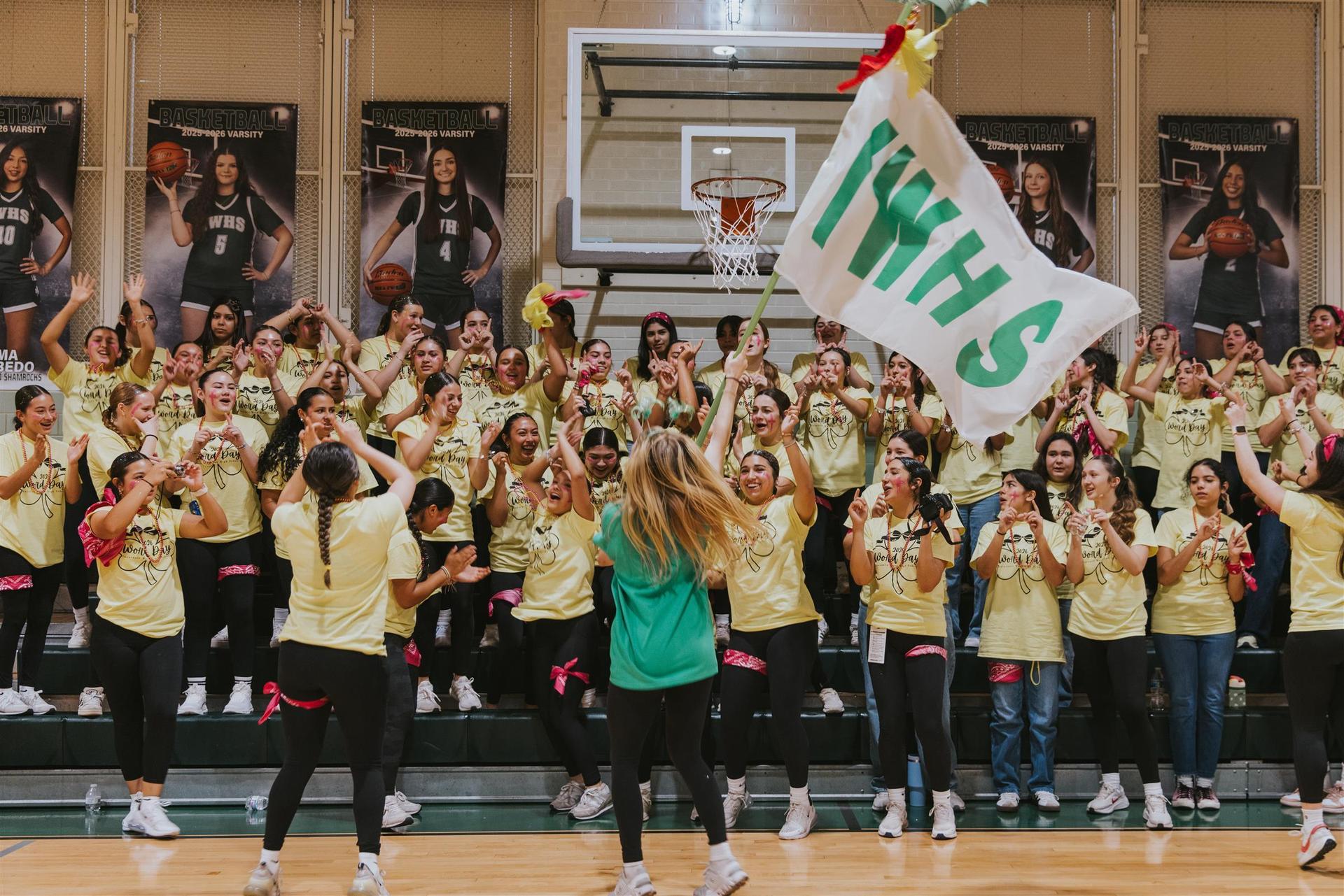 Cheering crowd in yellow shirts with a large flag reading 'WHS' in a gymnasium.