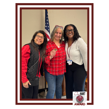 Three women standing together, one holding an award with a flag in the background.