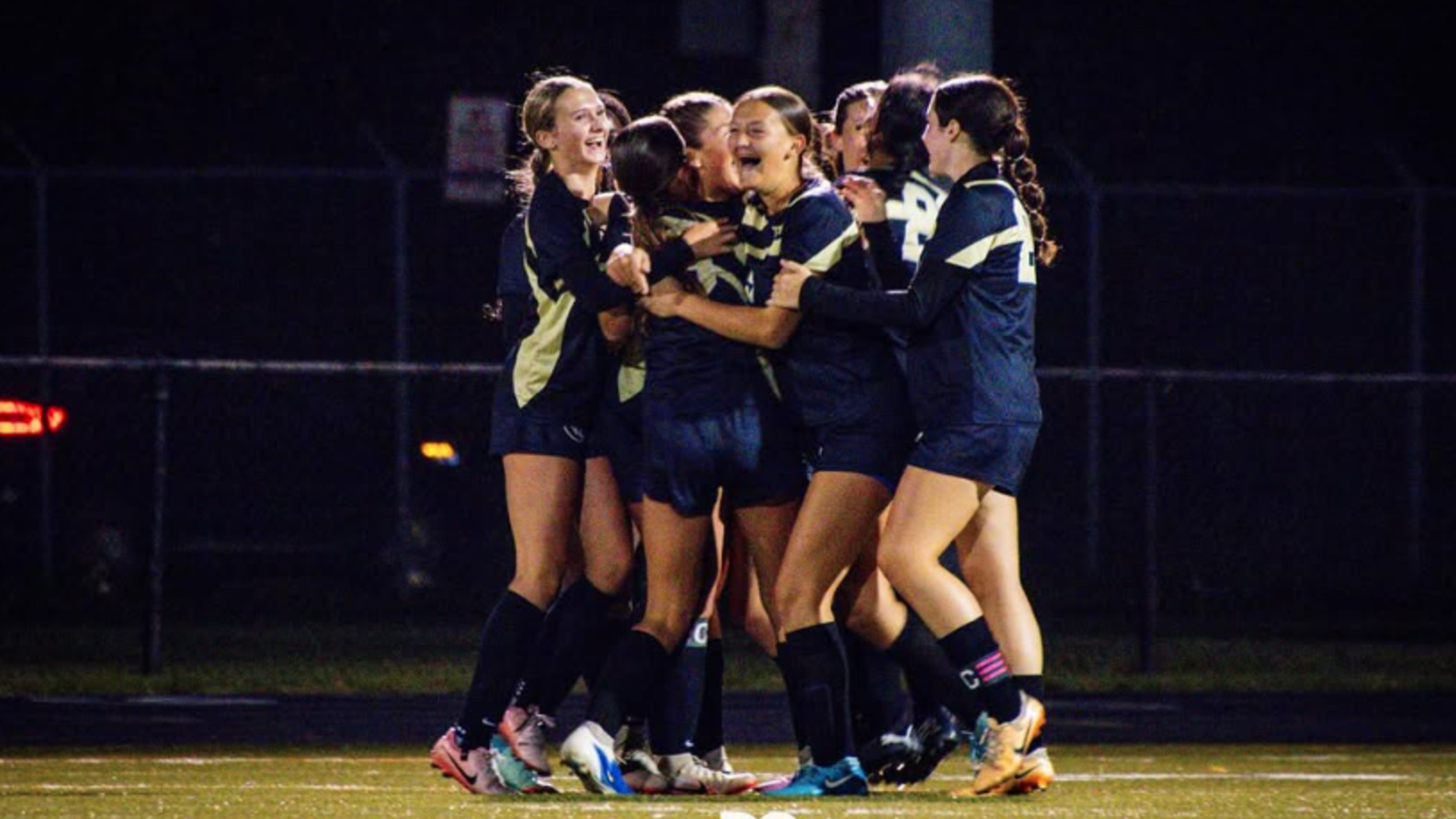 A group of female high school soccer players in dark uniforms hug and celebrate together on a field at night.