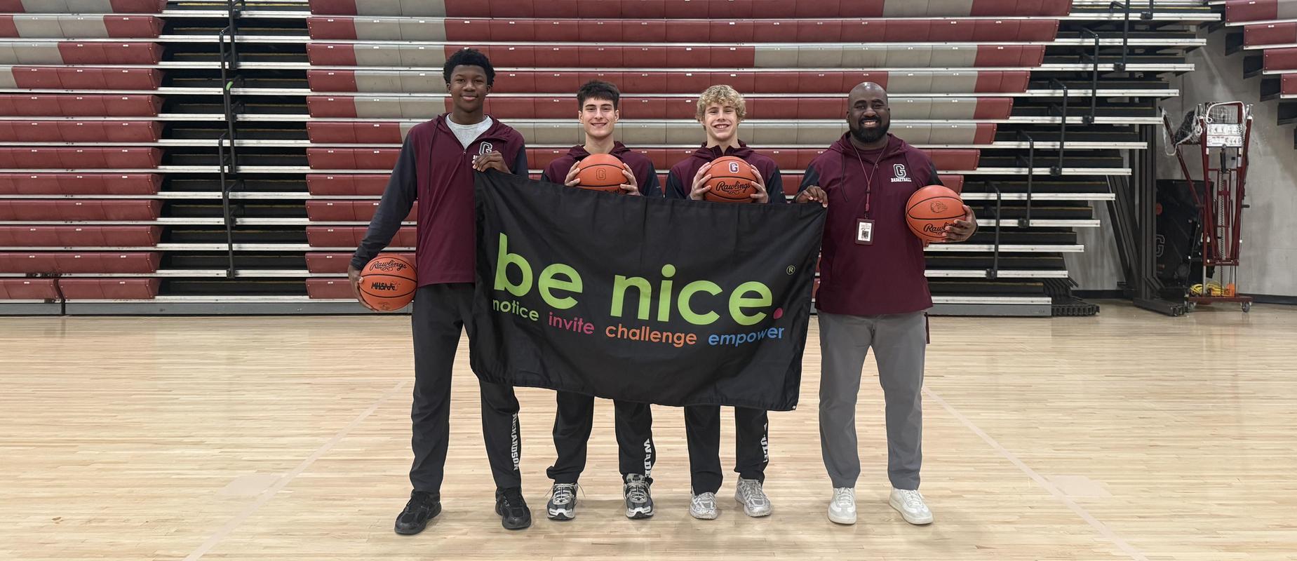three basketball players and the coach stand in the gym smiling with a be nice banner