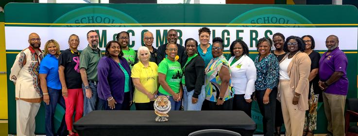 A diverse group of 20 individuals standing together in front of a school banner.