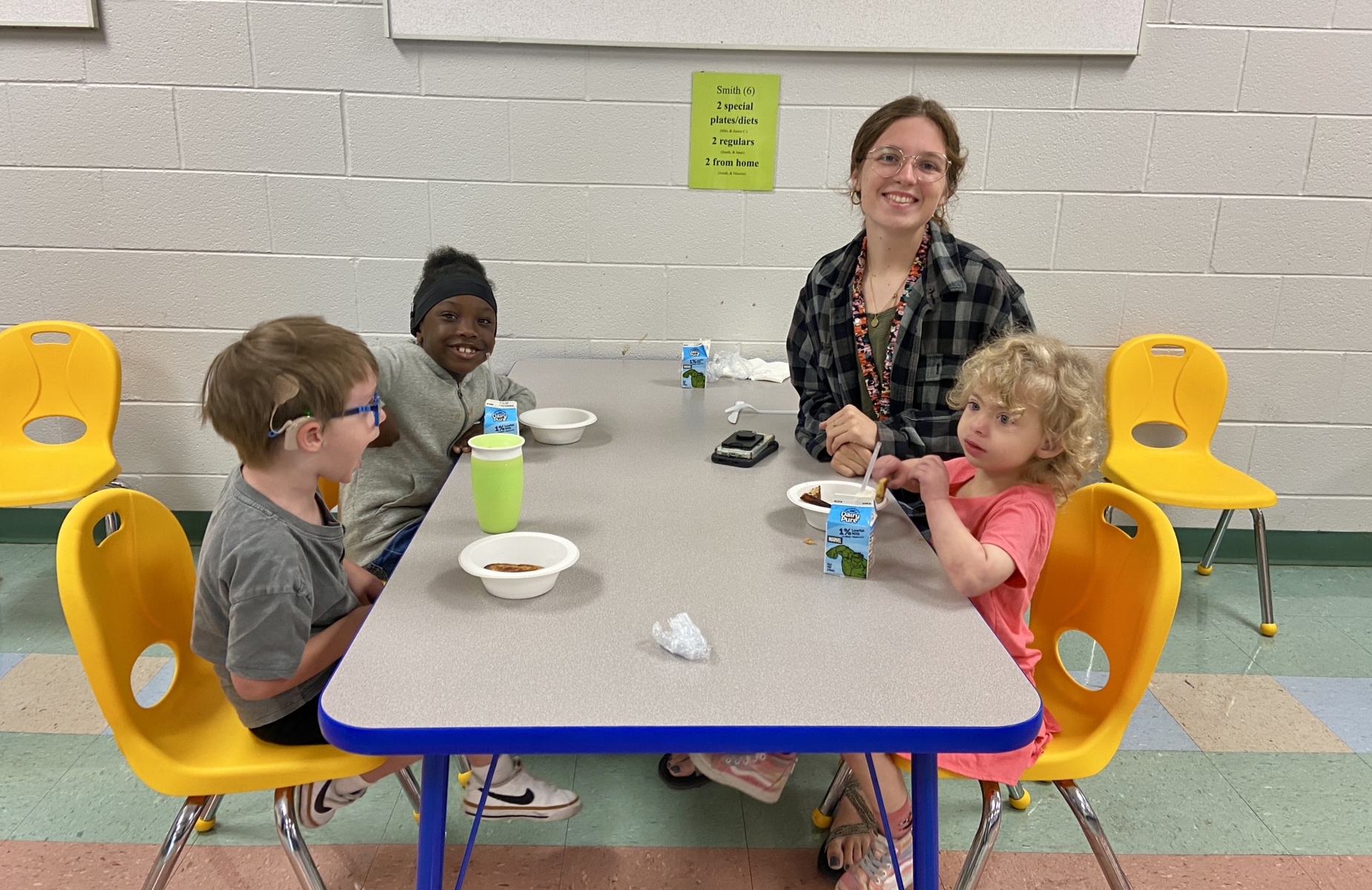 A group of four children enjoying a meal with an adult in a brightly colored cafeteria.