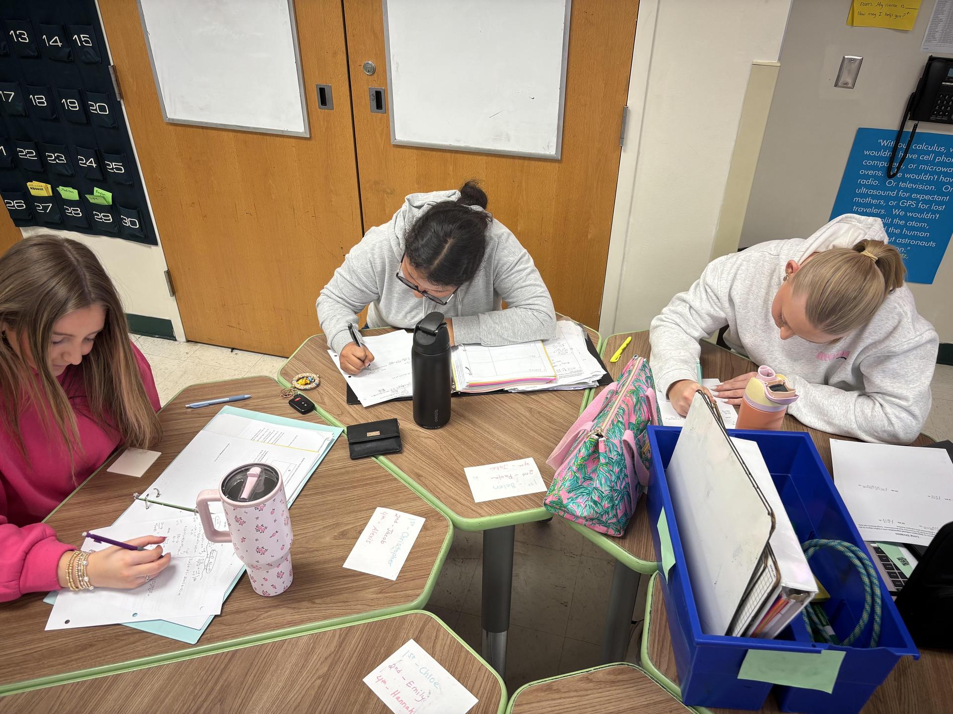 Three students writing notes at a table in a classroom, with supplies around them.
