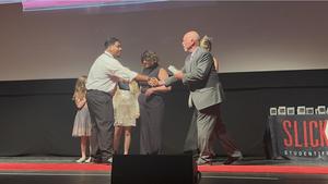 Ramses Gomez shakes hands with an award presenter on stage at the Slick Rock Student Film Festival, holding his plaque, while other presenters and children stand in the background.