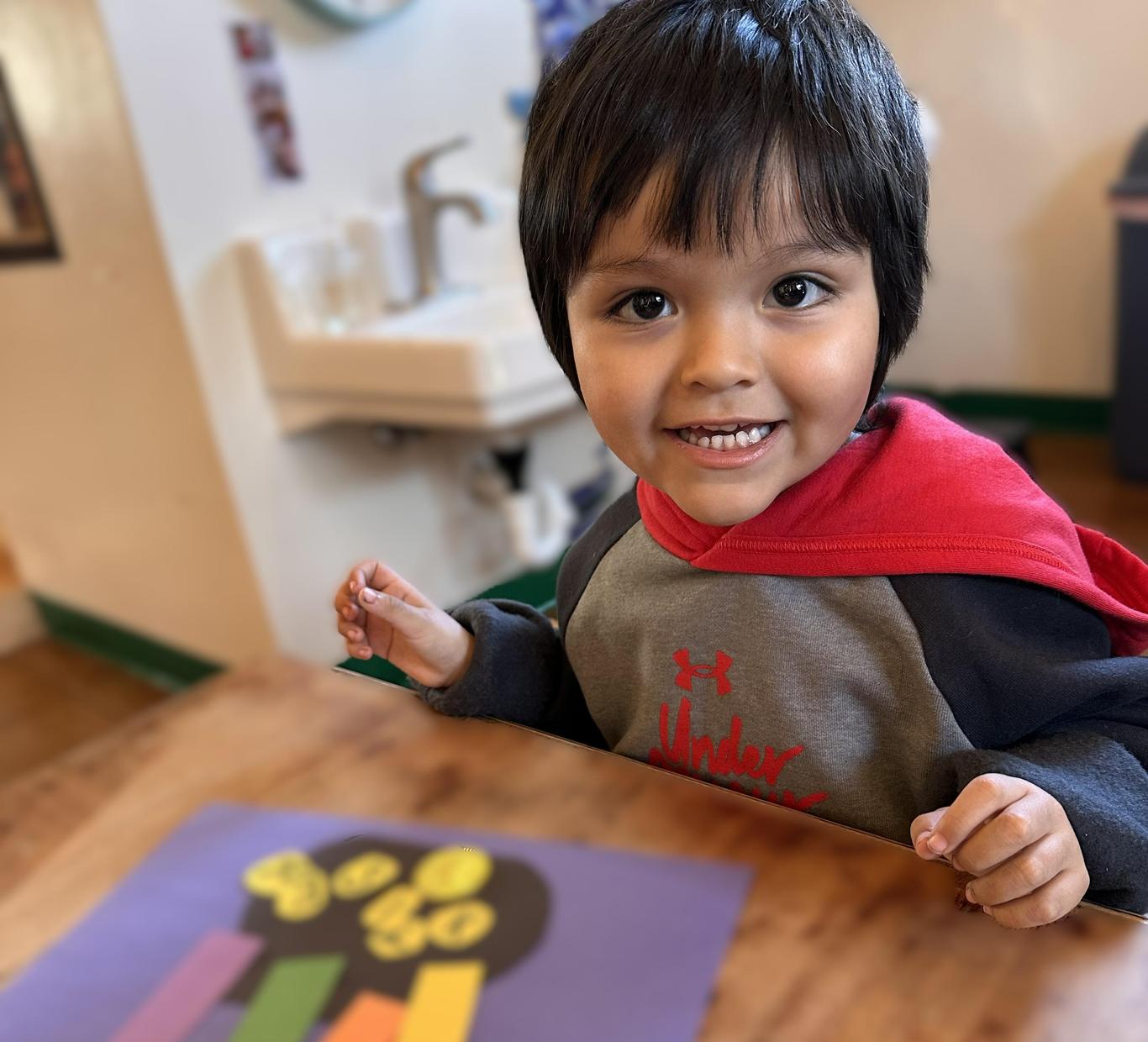 Smiling child with dark hair in a red hoodie, seated at a table with colorful artwork.