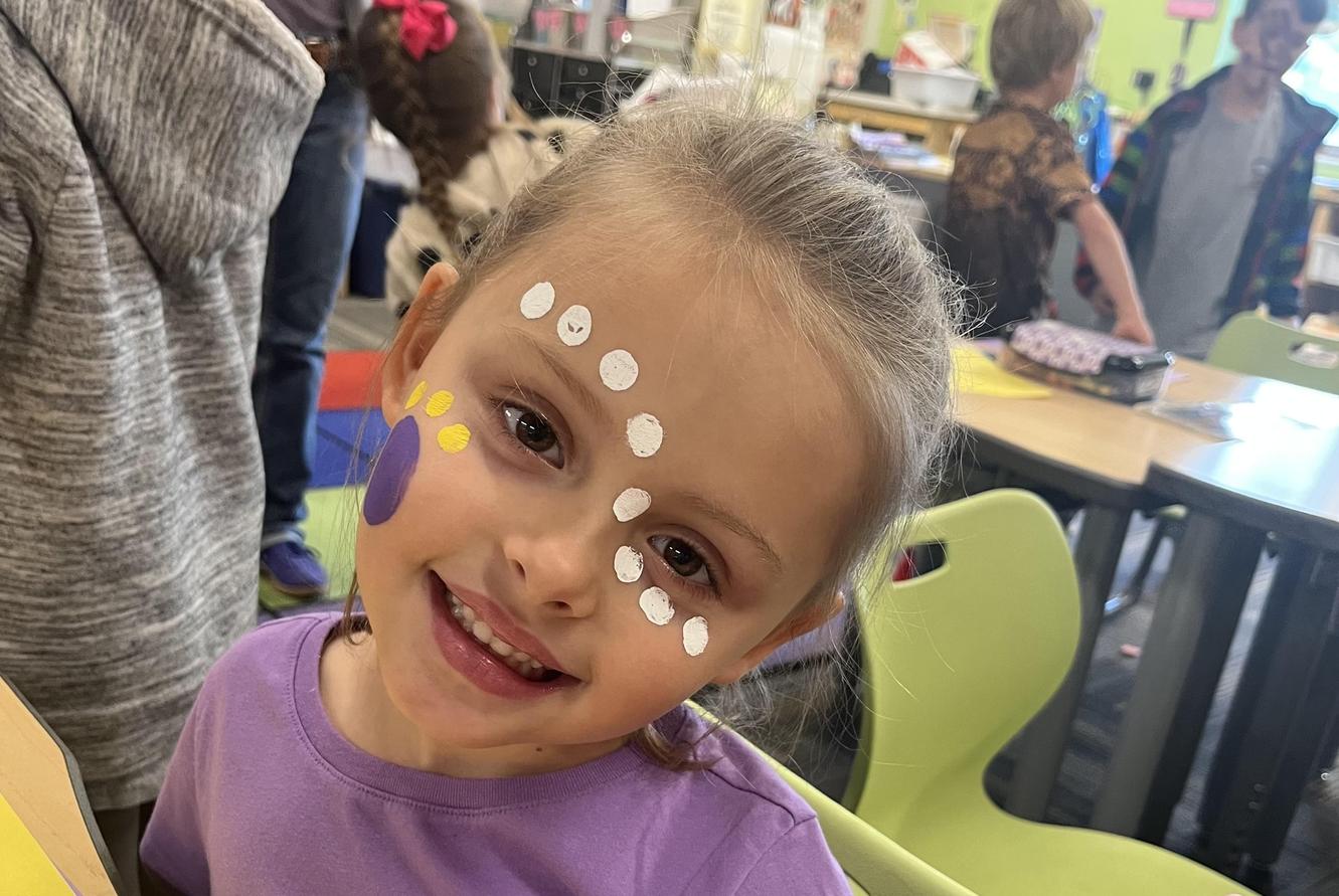 Smiling girl with colorful face paint poses at a classroom table.