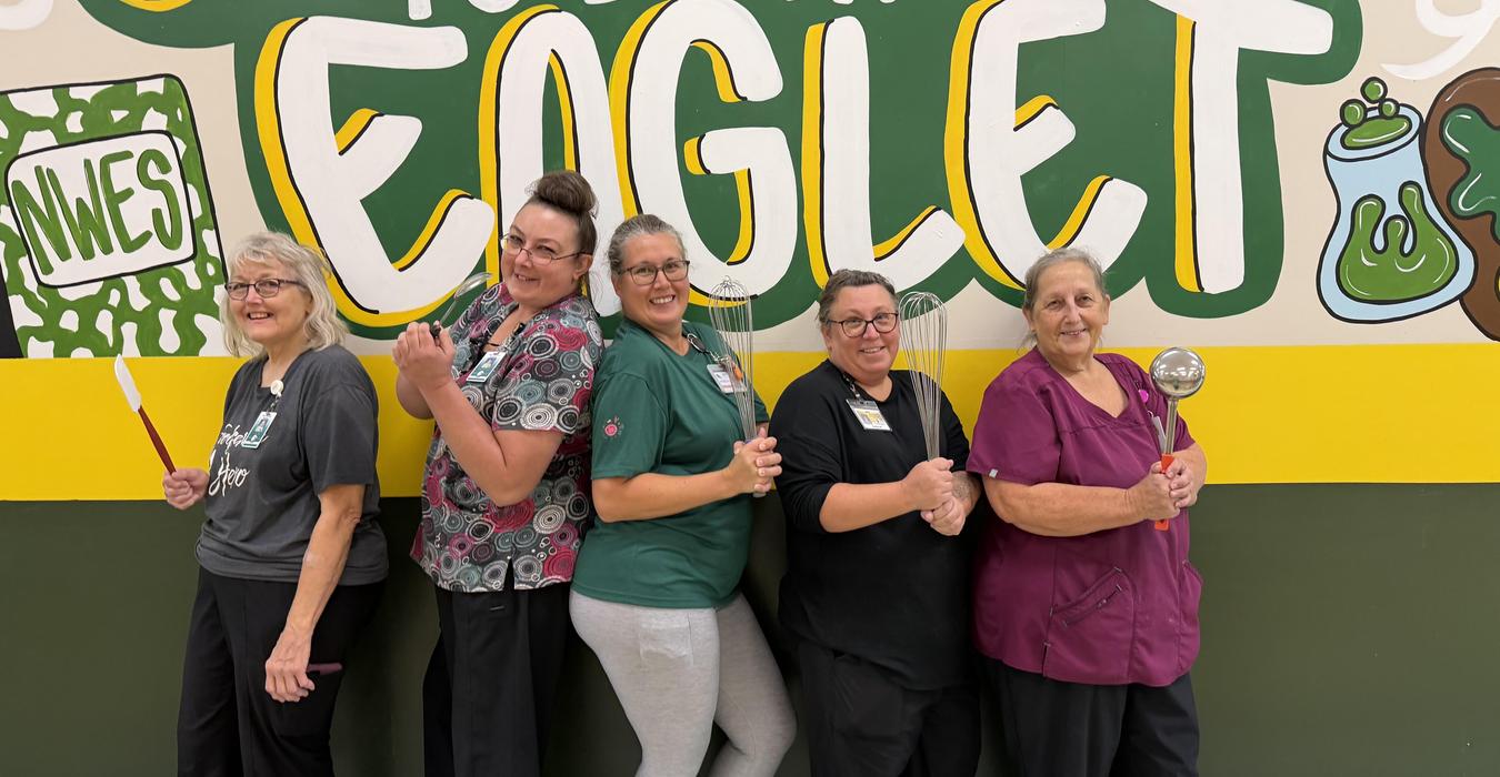 Five women stand together, smiling in front of a colorful mural featuring the word 'EGGLET'.