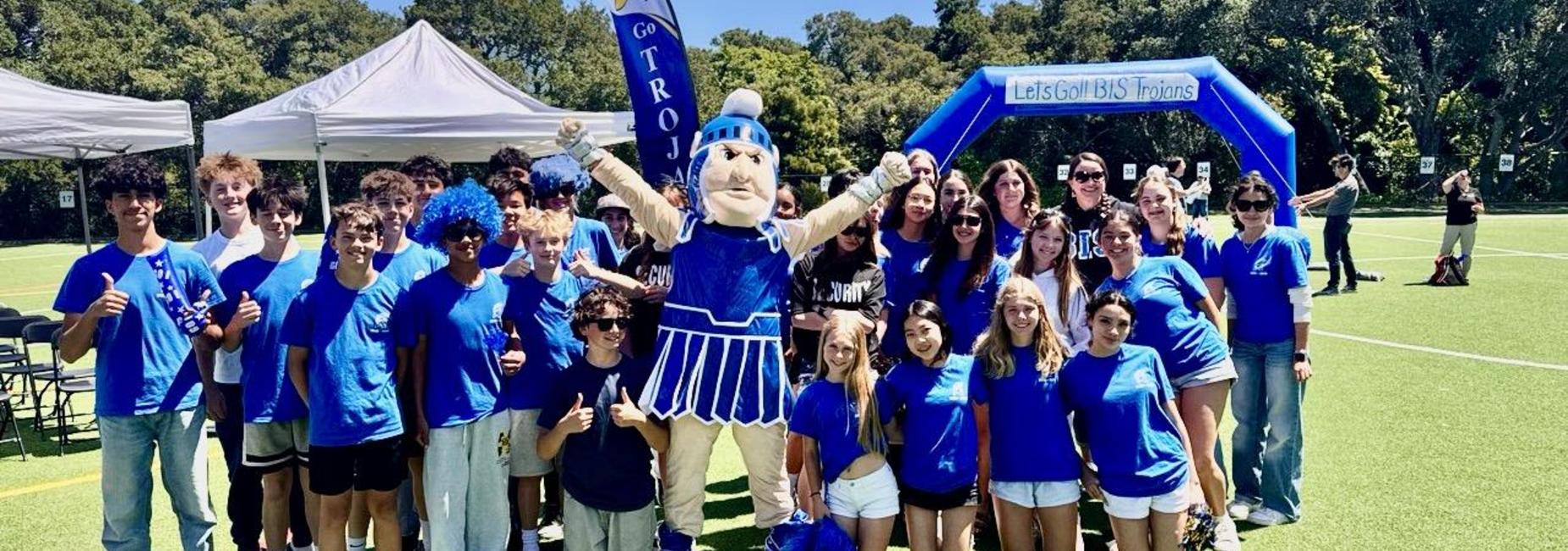 A cheerful group of BIS students wearing blue, posing with a mascot at a sports event.