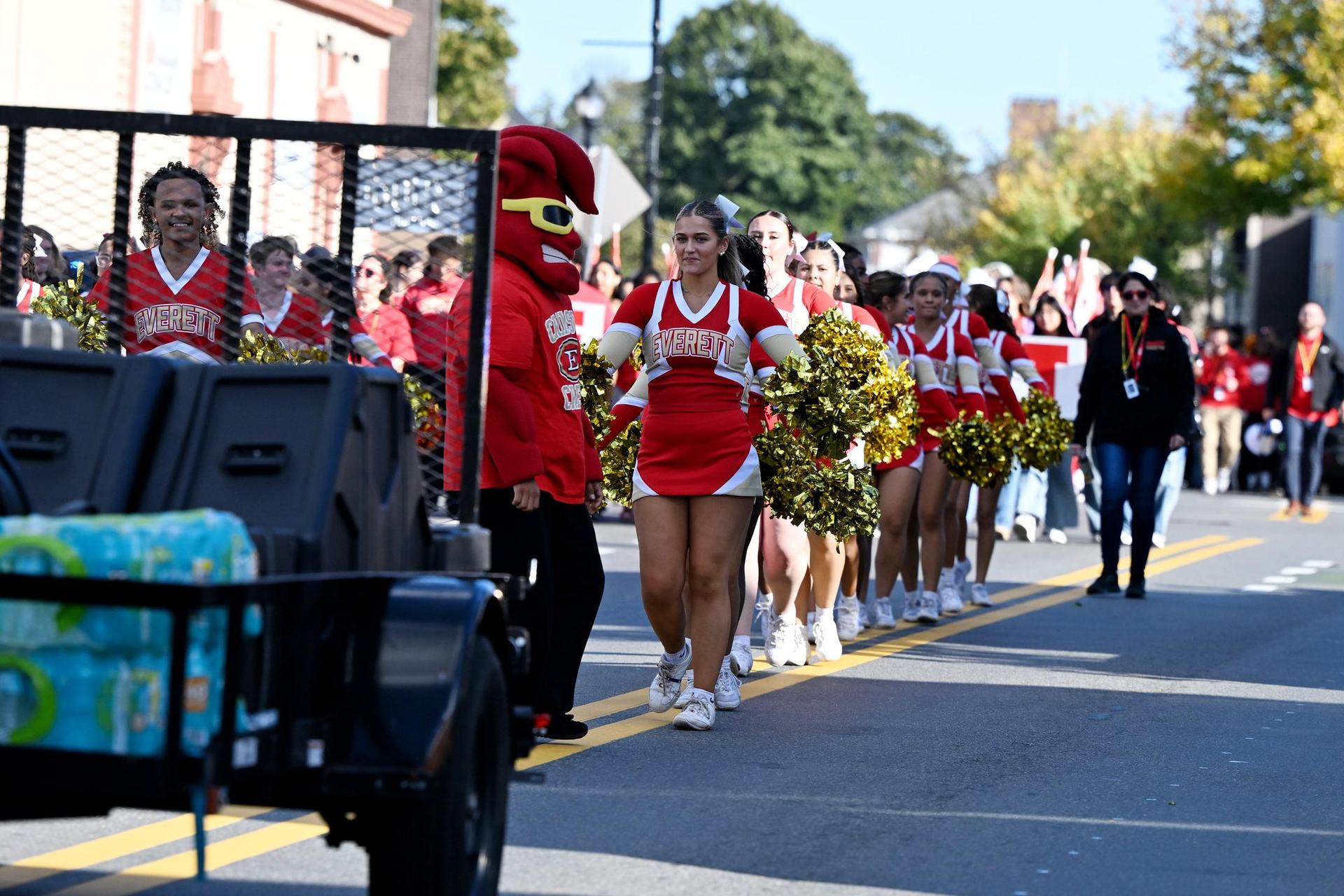 Cheerleaders and a mascot march in a parade on a sunny street.