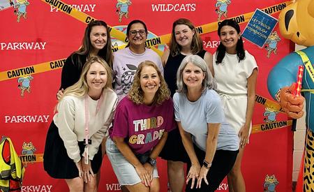 Seven women pose for a group photo in front of a red backdrop with caution tape and a cat mascot.