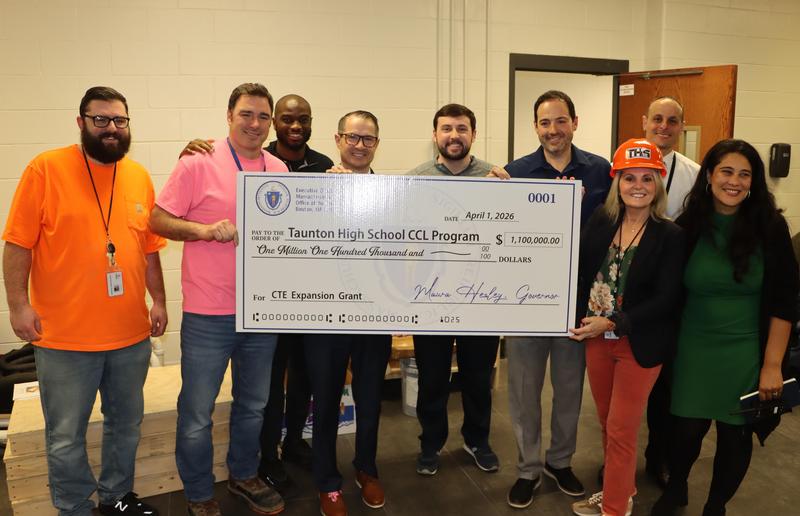 A group of adults stand together indoors holding a large ceremonial check, with one woman wearing an orange THS construction hard hat.