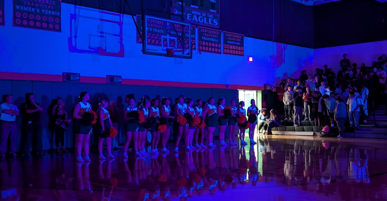 Cheerleaders in uniform stand in a gym under blue lighting, with a crowd in the background.