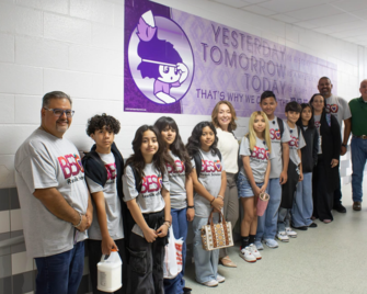 A group of students and adults pose together in a hallway, wearing matching t-shirts.