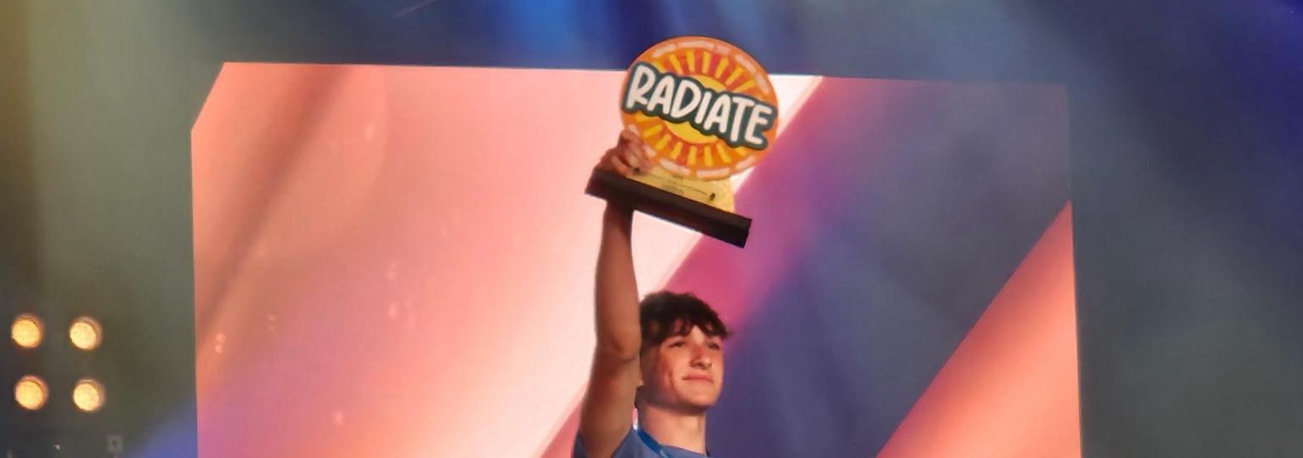 Teenage boy raising his arm in celebration on stage with two others, one in formal attire.