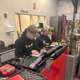 Two students working together on electronics with tools and documents.
