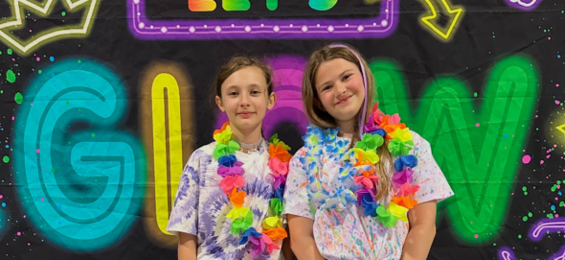 Two girls wearing colorful leis and party outfits stand in front of a neon backdrop.