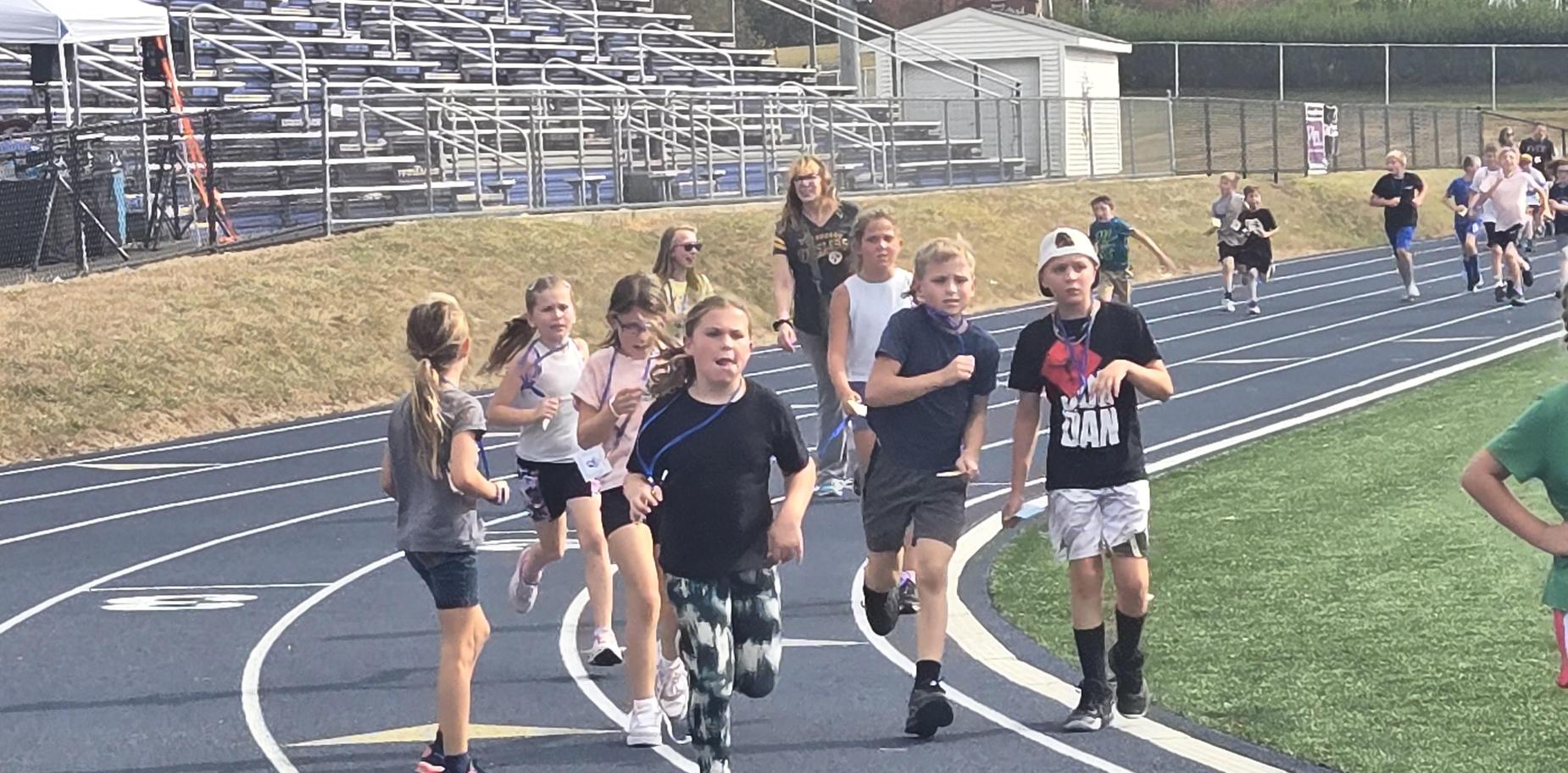 A group of children running on a track, some wearing athletic gear, with excited expressions.
