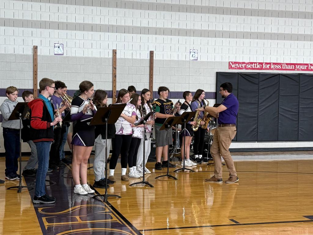 Students in the band playing instruments in the gym with their teacher directing them.