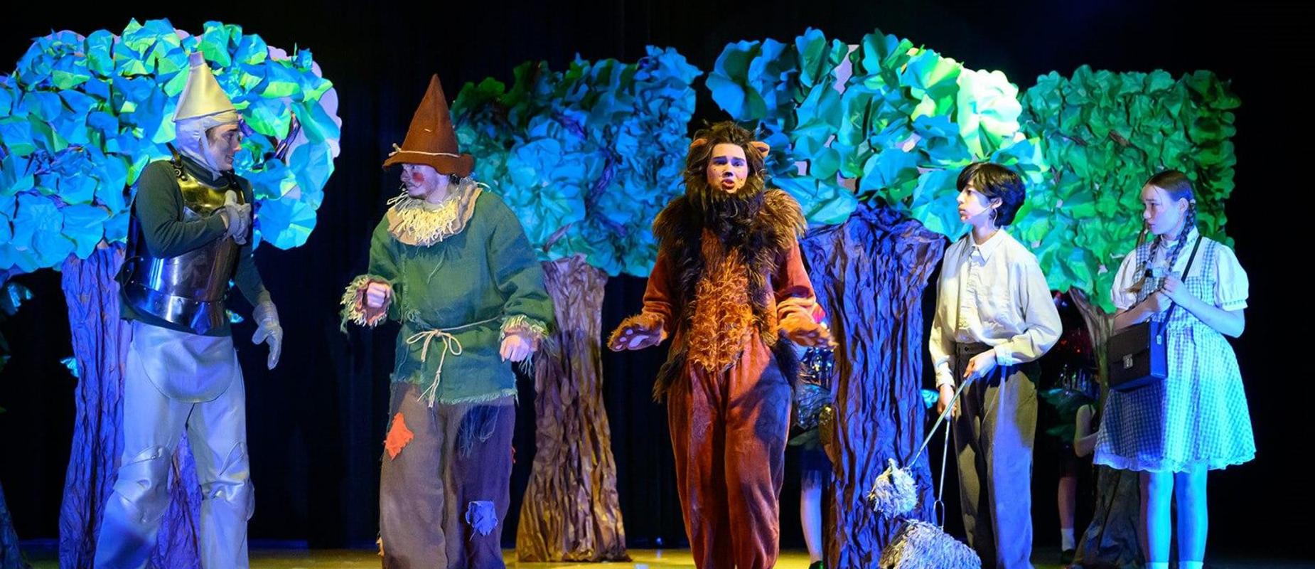 Tin Man, Scarecrow, Lion, Toto, and Dorothy stand in front of trees made from paper on stage