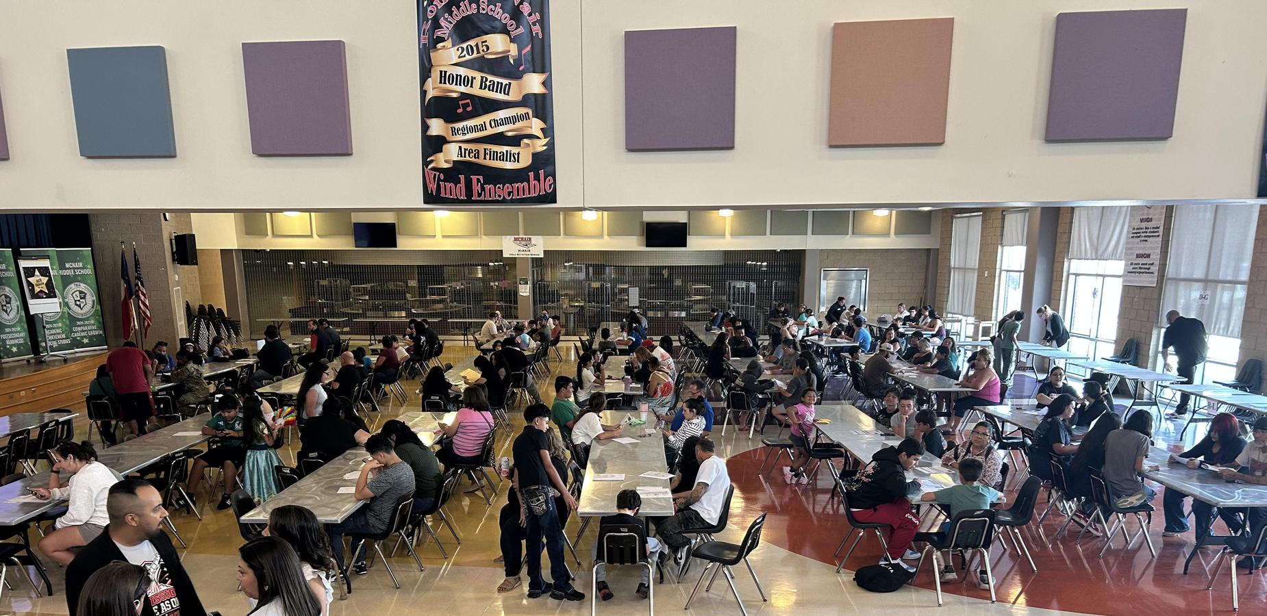 A large indoor space filled with people seated at tables, engaging in various activities.