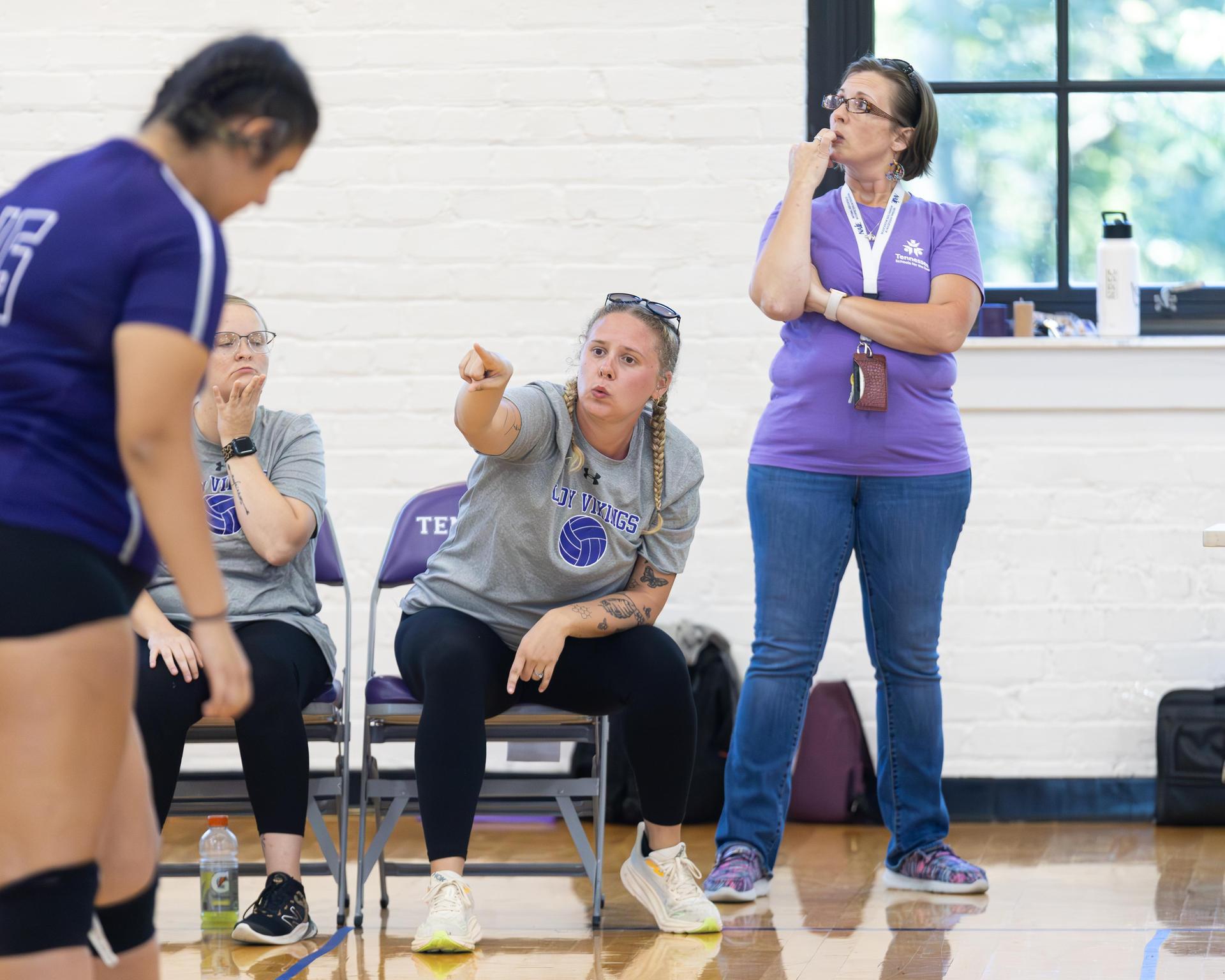 A coach points toward a player while another coach observes, both in gray shirts, during a volleyball match.