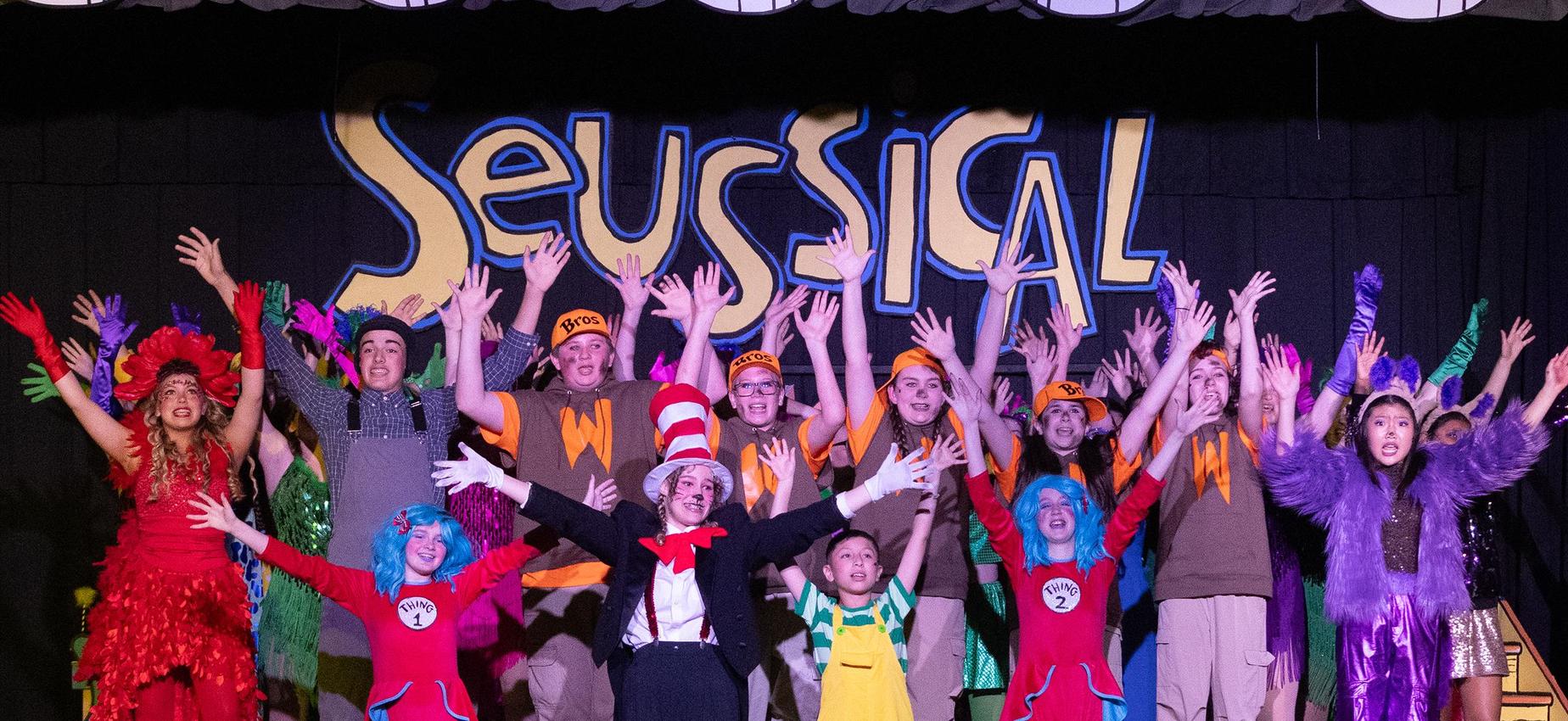 A lively theater performance with actors in colorful costumes and raised hands.