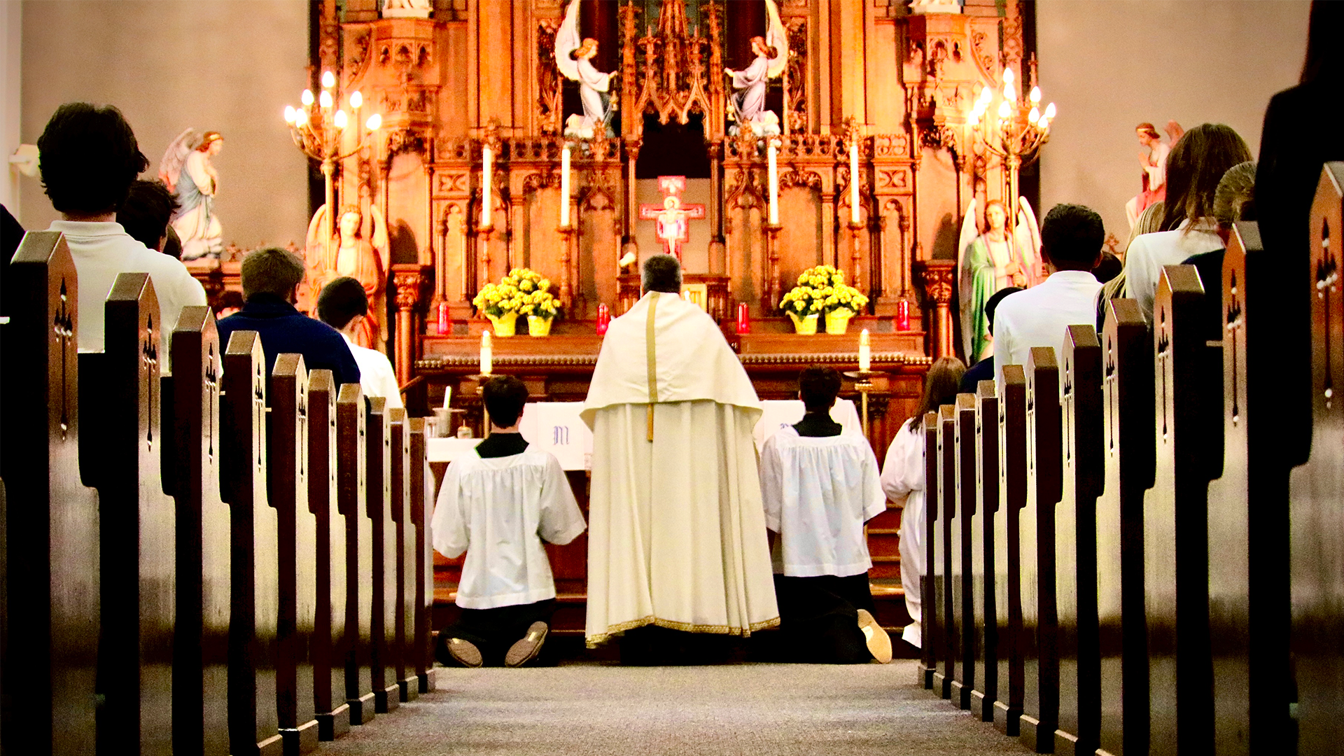 priest in white vestments faces altar with two altar boys genuflecting
