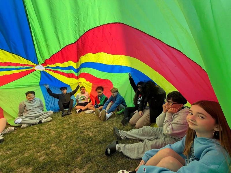 Group of children sitting under a colorful parachute on the grass.