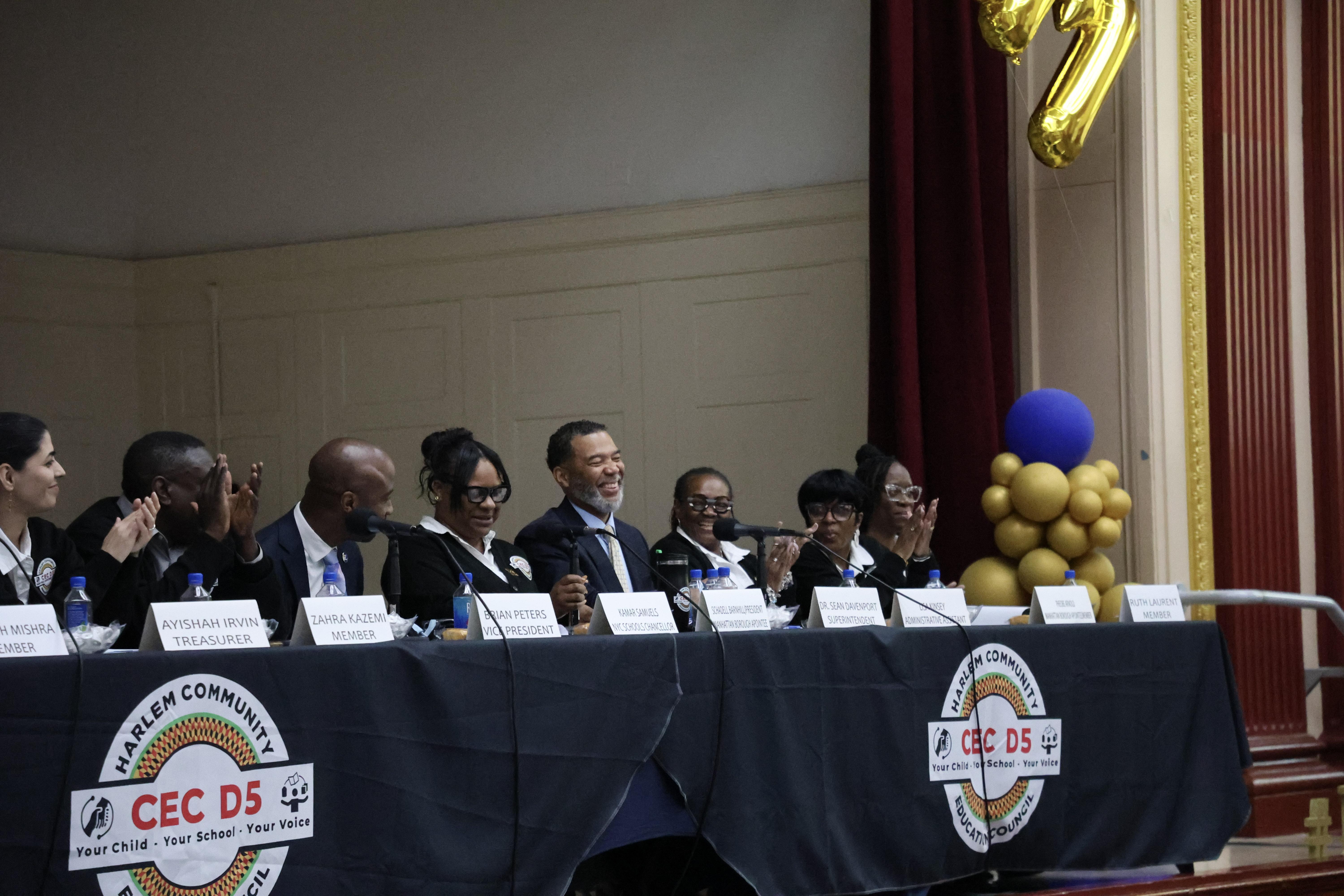 Panel of speakers at a community meeting with decorative balloons.