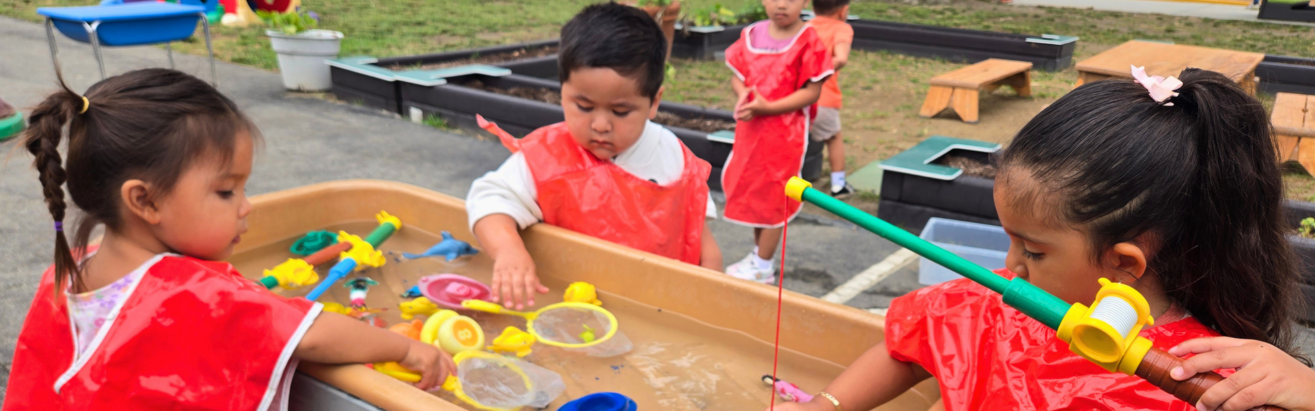 Young children in red aprons playing with water and toys in an outdoor activity area.